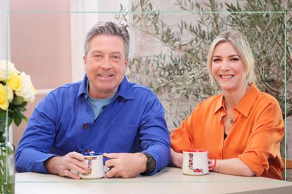 John Torode (left) and Lisa Faulkner (right) sitting at a table with a mug each