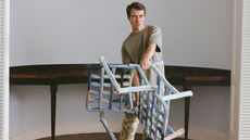 A young man dressed in a pale, dark green t-shirt and camouflaged trousers holds a chair made from what looks like soft colorful marshmallows.