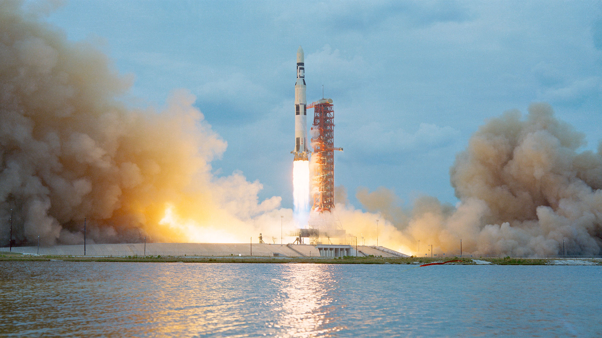 A large white rocket with black stripes lifts off into a blue sky besides a red launch tower.