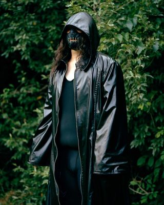 Photograph of person wearing mask and black coat against natural green backdrop