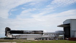 a white spacex falcon heavy rocket heads out of its hangar in a horizontal configuration under a blue sky.