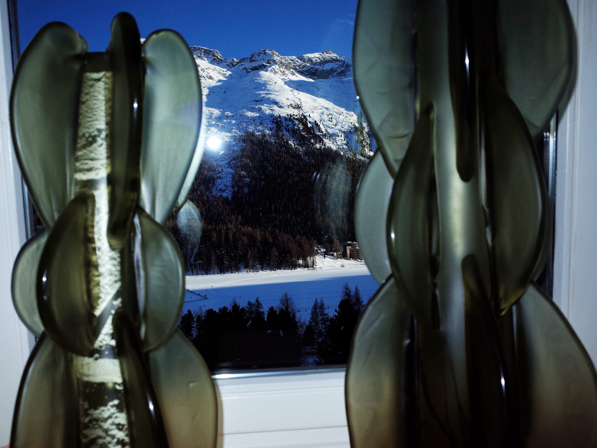 An alpine view caught from a window indoors between two multi-part glass sculptures.