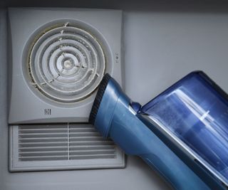 Cleaning wall-mounted dusty hood ventilation grill in bathroom with vacuum cleaner.