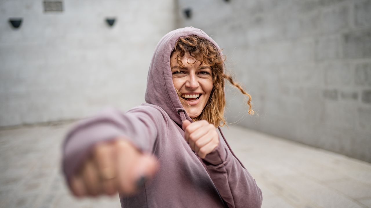 Woman shadowboxing, throwing a jab at the camera. She is wearing a lilac hoodie with the hood up.