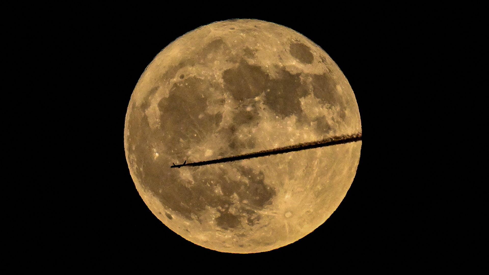A passenger airline is pictured silhouetted leaving a thick exhaust trail while travelling across the against the yellow disk of the full 'Flower Moon' on May 12, 2025.