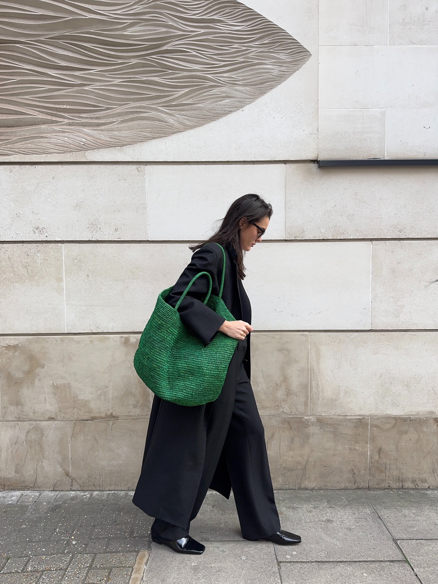 Valeria Soba wears a black coat, trousers and shoes, plus a large green raffia tote bag.