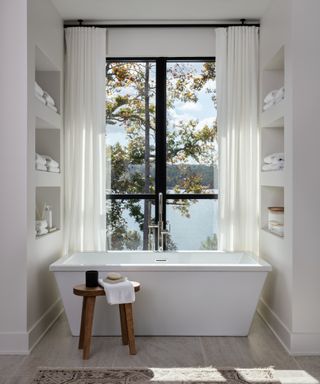 white bathroom with custom shelving each side of bath with towels, wooden stool, rug, view outside