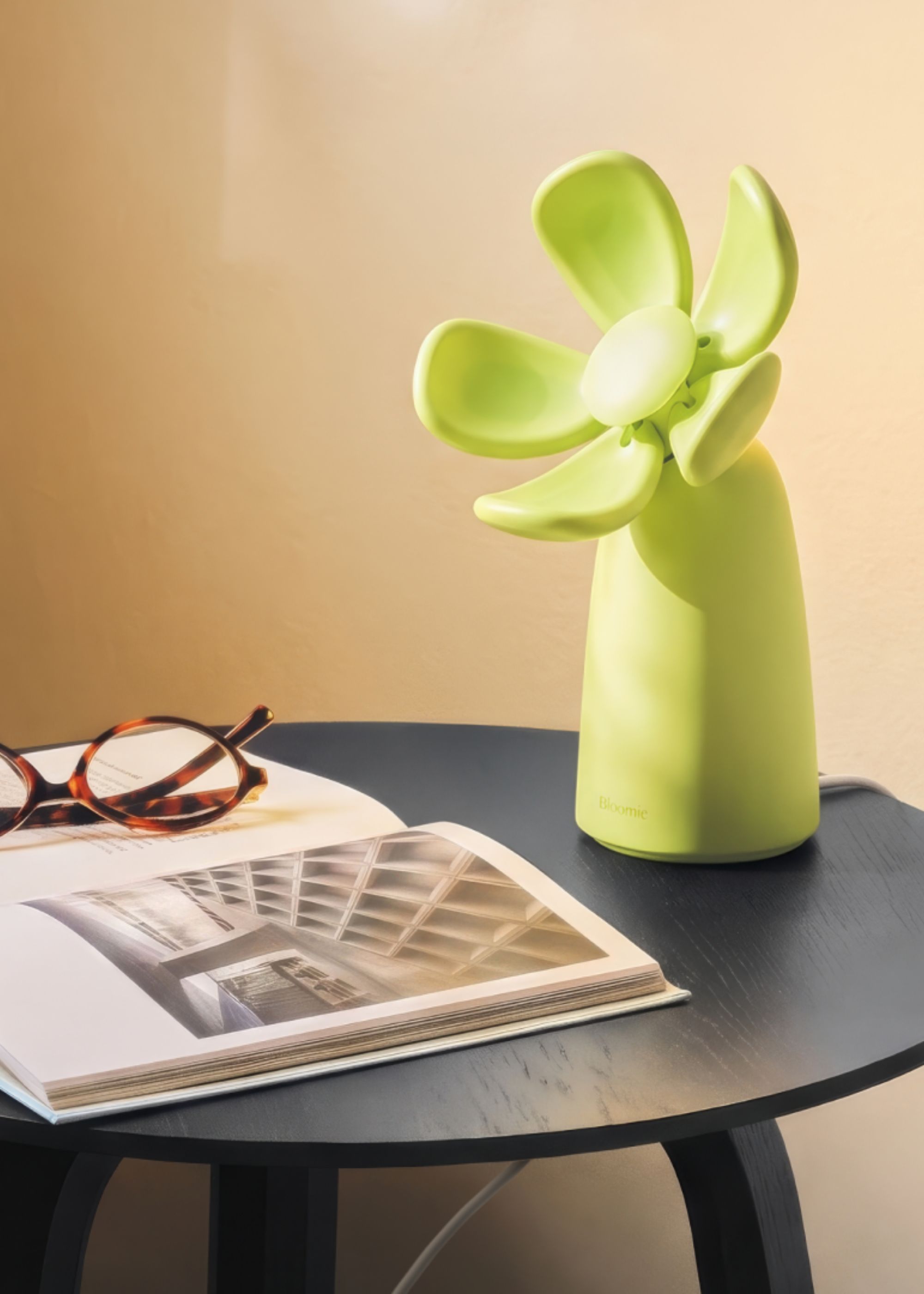 A lime green flower-shaped air quality checker from Bloomie on a dark blue side table with an open book and tortoise-shell glasses