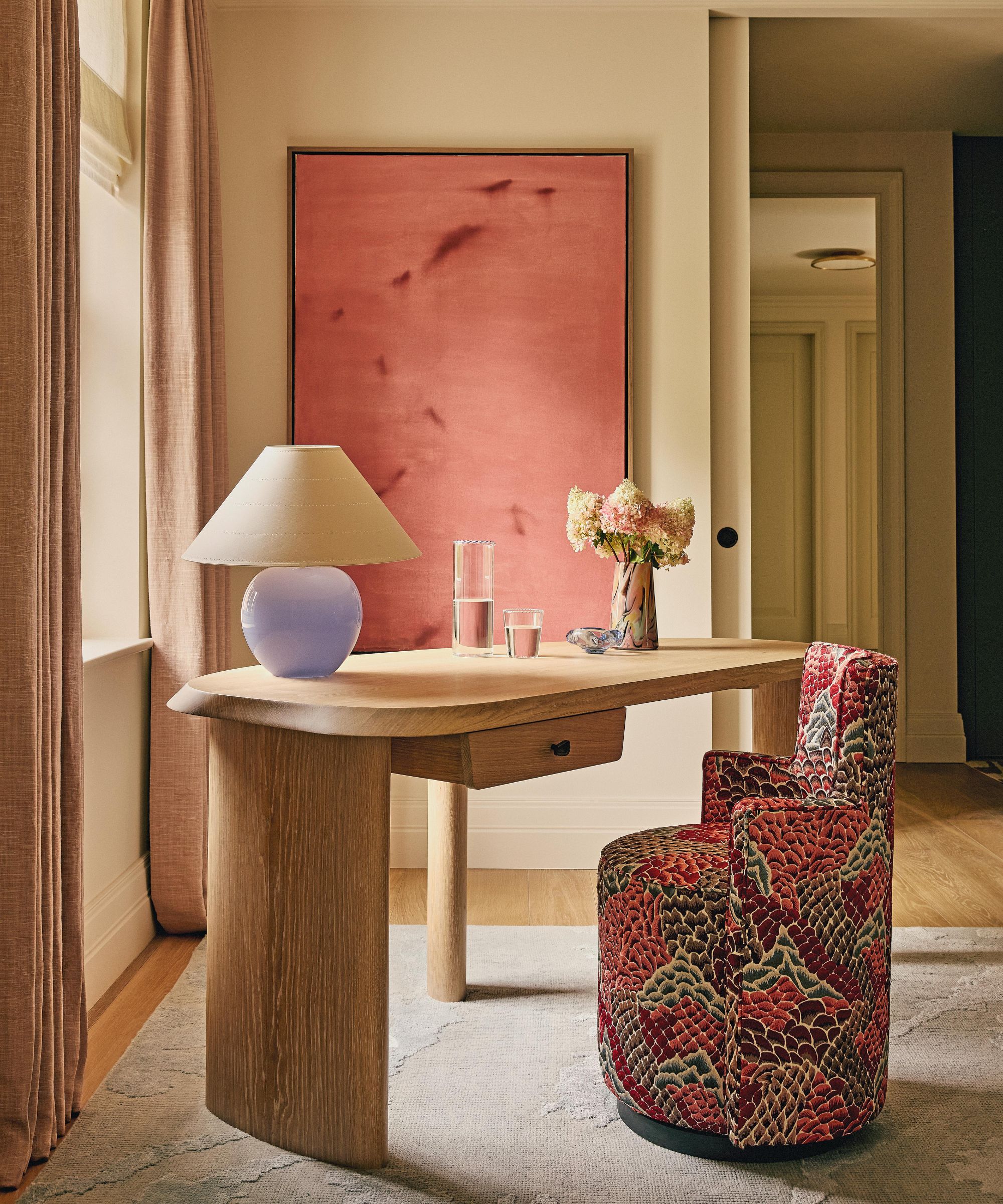 a pale pink bedroom corner with a large pink canvas. an oak desk sits at an angle with an upholstered vanity chair