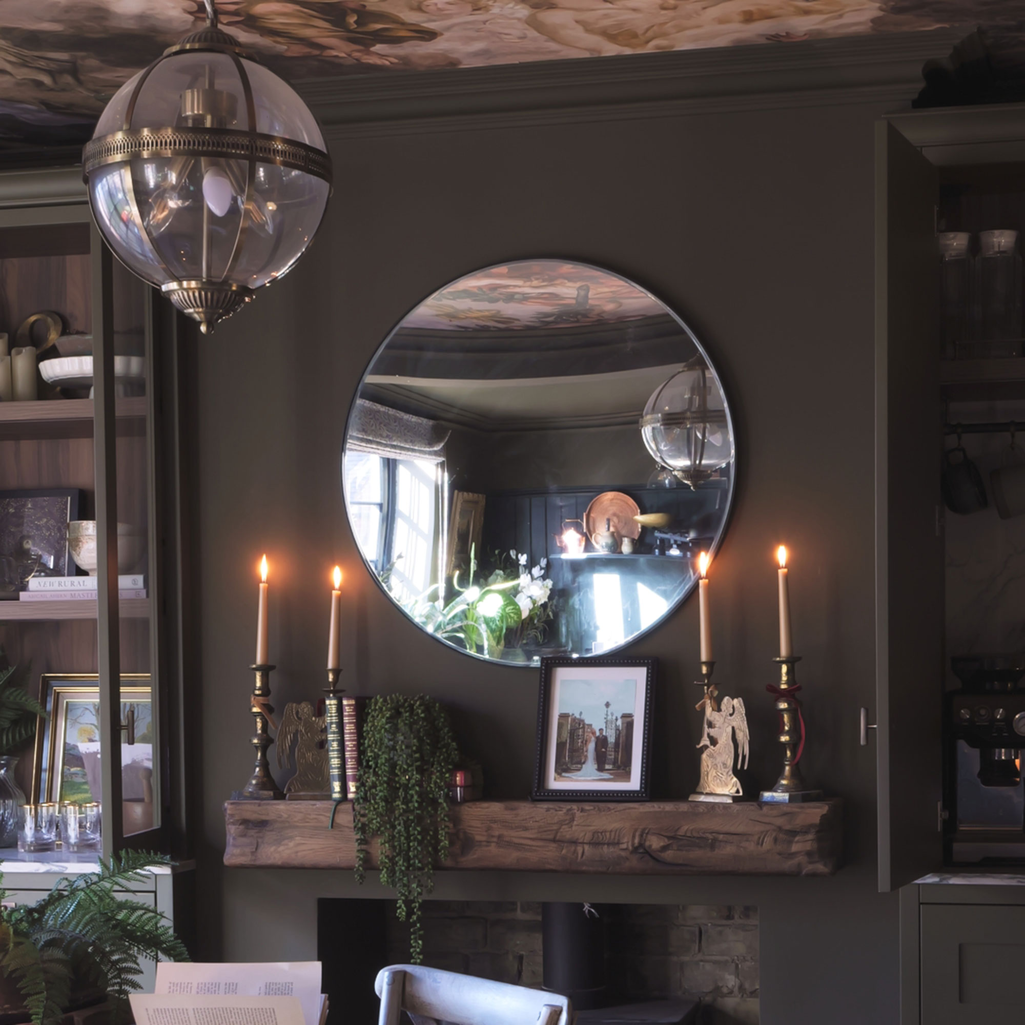 Dark grey-green room with candles on wooden mantlepiece below circular mirror