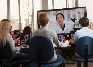 Students with digital tablets watching instructor on monitor -