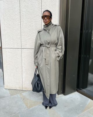 A woman wears a trench coat, with gray trousers, black pointed toe heels, and a black tote bag.