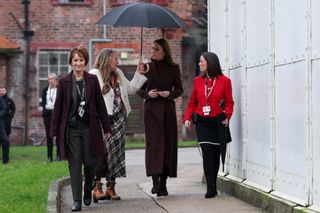 Three women walking outside a prison next to Kate Middleton, with one holding an umbrella over the princess, who is wearing a long maroon plaid coat and black boots