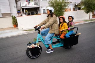 A woman rides blue Trek Fetch cargo bike with two children on the back and toys in the front baskets