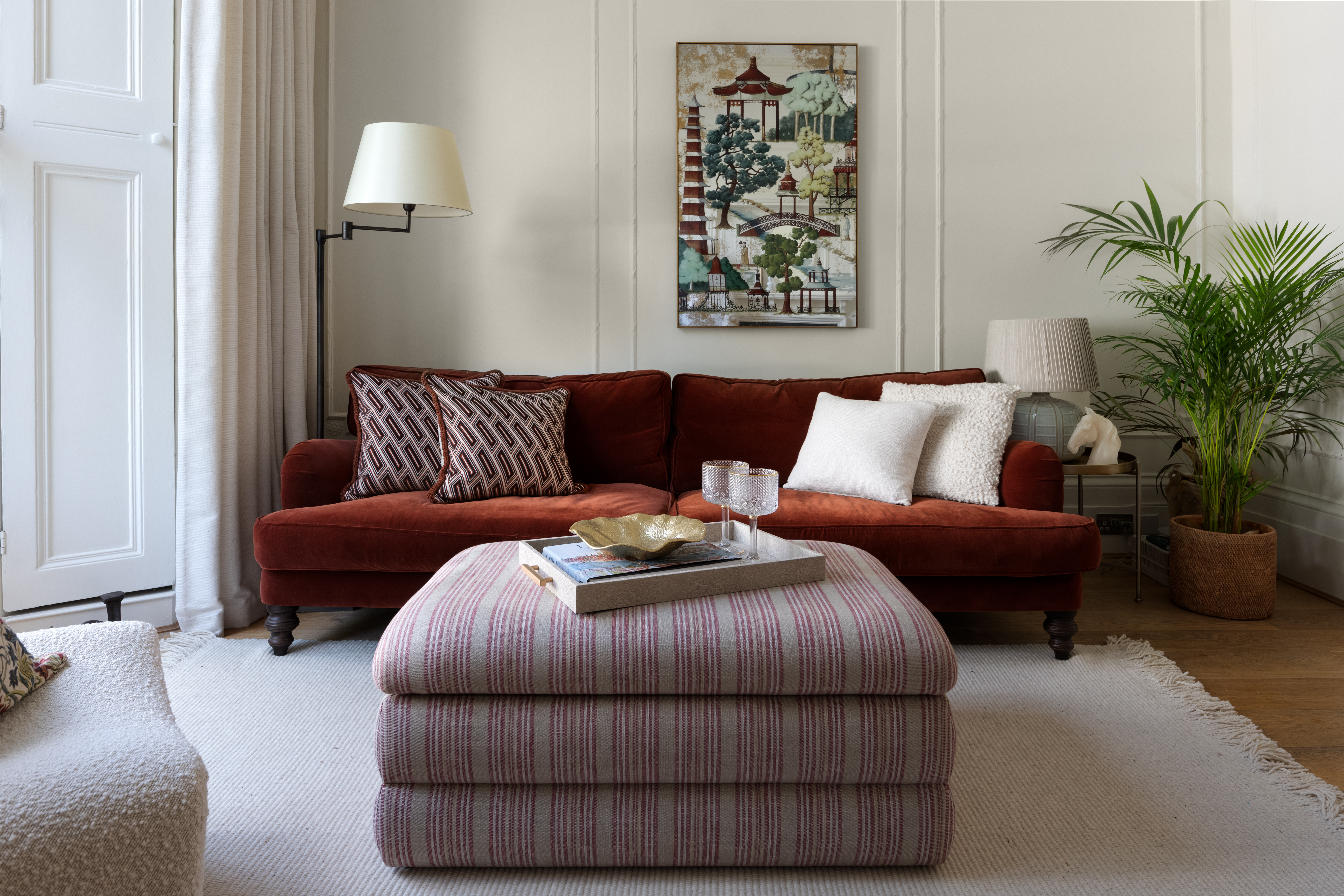 A living room with a modern vibe, a red velvet sofa and a striped ottoman. Wainscotting on the wall.