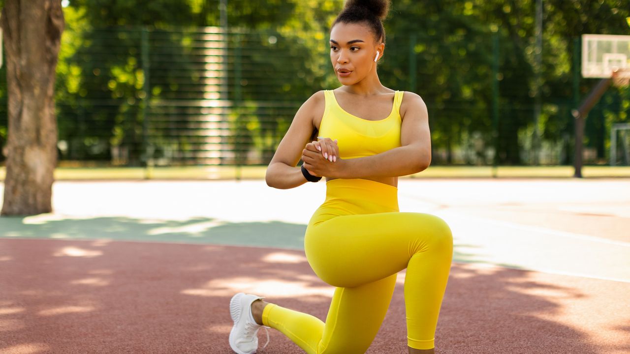 A woman concentrates as she performs a lunge with rotation exercise. She is outside on a basketball court, dressed in workout gear, and there are leafy trees in the distance.