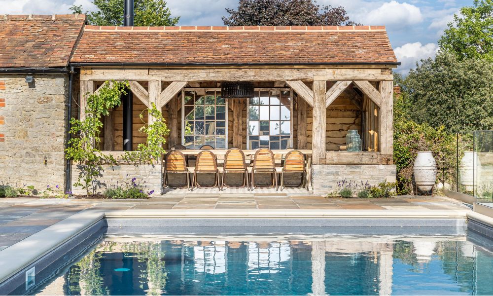 A rustic open-sided oak frame pool house with a clay tile roof, antique glazing and a long teak dining table, beside a slate-edged pool with stone flags and lavender planting.