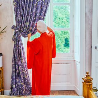 Angel from Escape to the Chateau in a orange outfit, facing the large window touching the full length heavy purple floral curtains