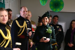 Prince William wearing a military uniform holding a Guinness standing next to Princess Kate in a room of soldiers with green decorations on St. Patrick's Day 2019