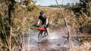 A white male bikepacker rides through a river crossing