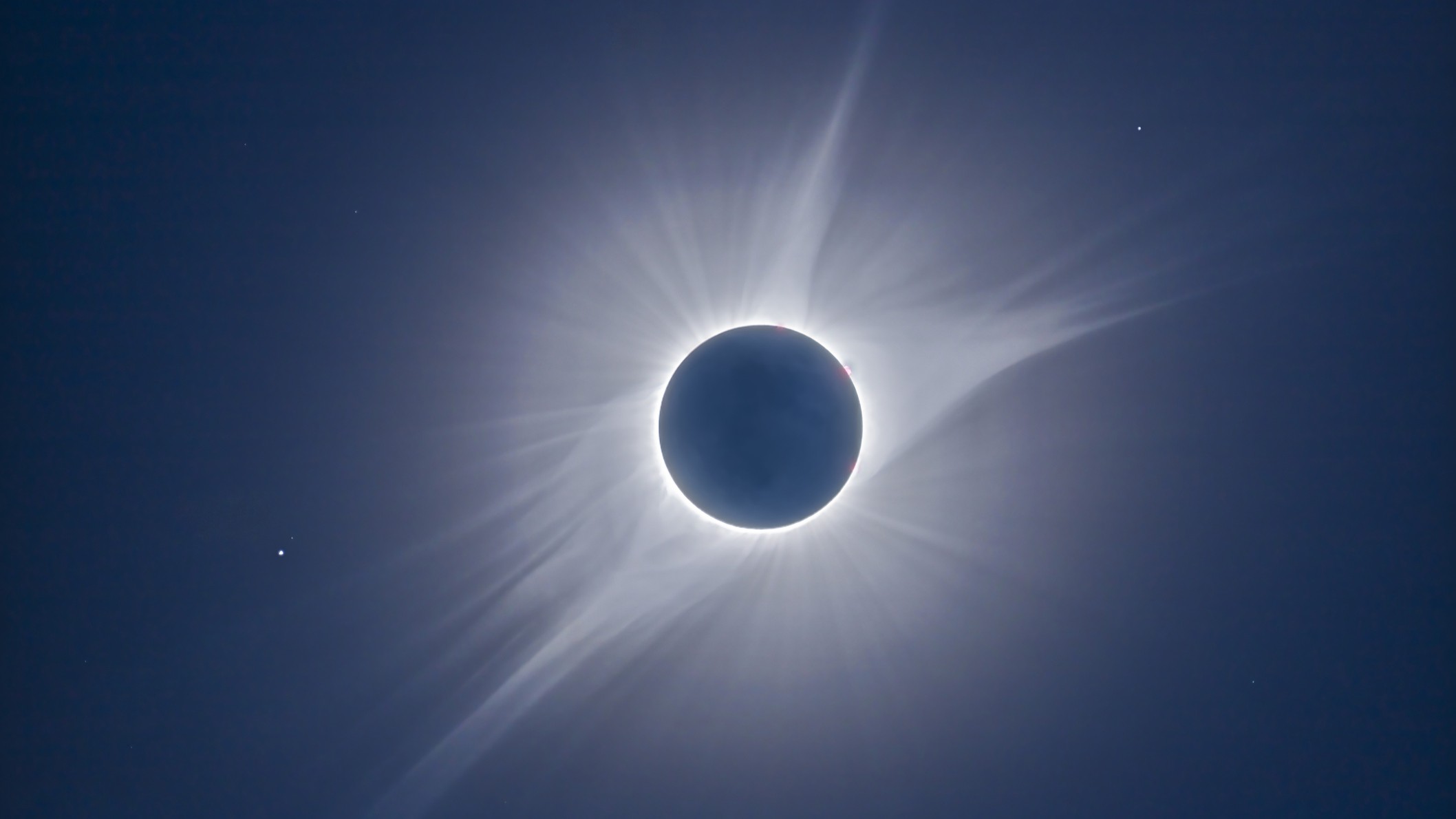 view of the sun during a total solar eclipse surrounded by a white halo with enormous white streamers firing off in all directions.