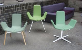 Outside daytime image, low brick wall, concrete floor, three different style chairs in various shades of pastel green, purple sofa in the background, bottom of bicycle wheels