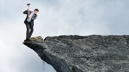 A businessman with a sledgehammer chips away at the part of a cliff he's standing on.