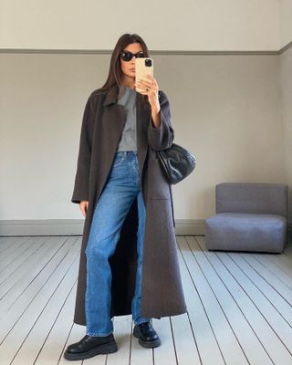 British influencer Marianne Smyth poses for a mirror selfie wearing Wayfarer sunglasses, a gray T-shirt, black leather bag, straight-leg jeans, and chunky black boots.