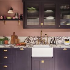a kitchen with pink walls and purple cabinetry with a marble splashback and countertop, brass fixtures and a Belfast sink