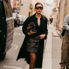 Woman wears mini skirt, black blouse, black trench coat, tights, and heels. 