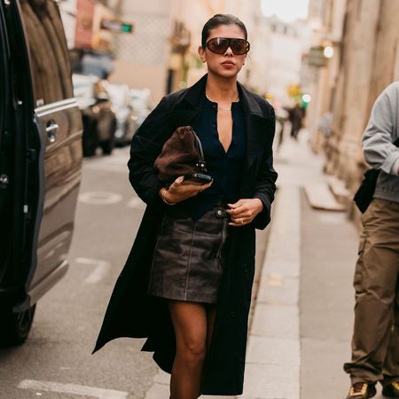 Woman wears mini skirt, black blouse, black trench coat, tights, and heels. 