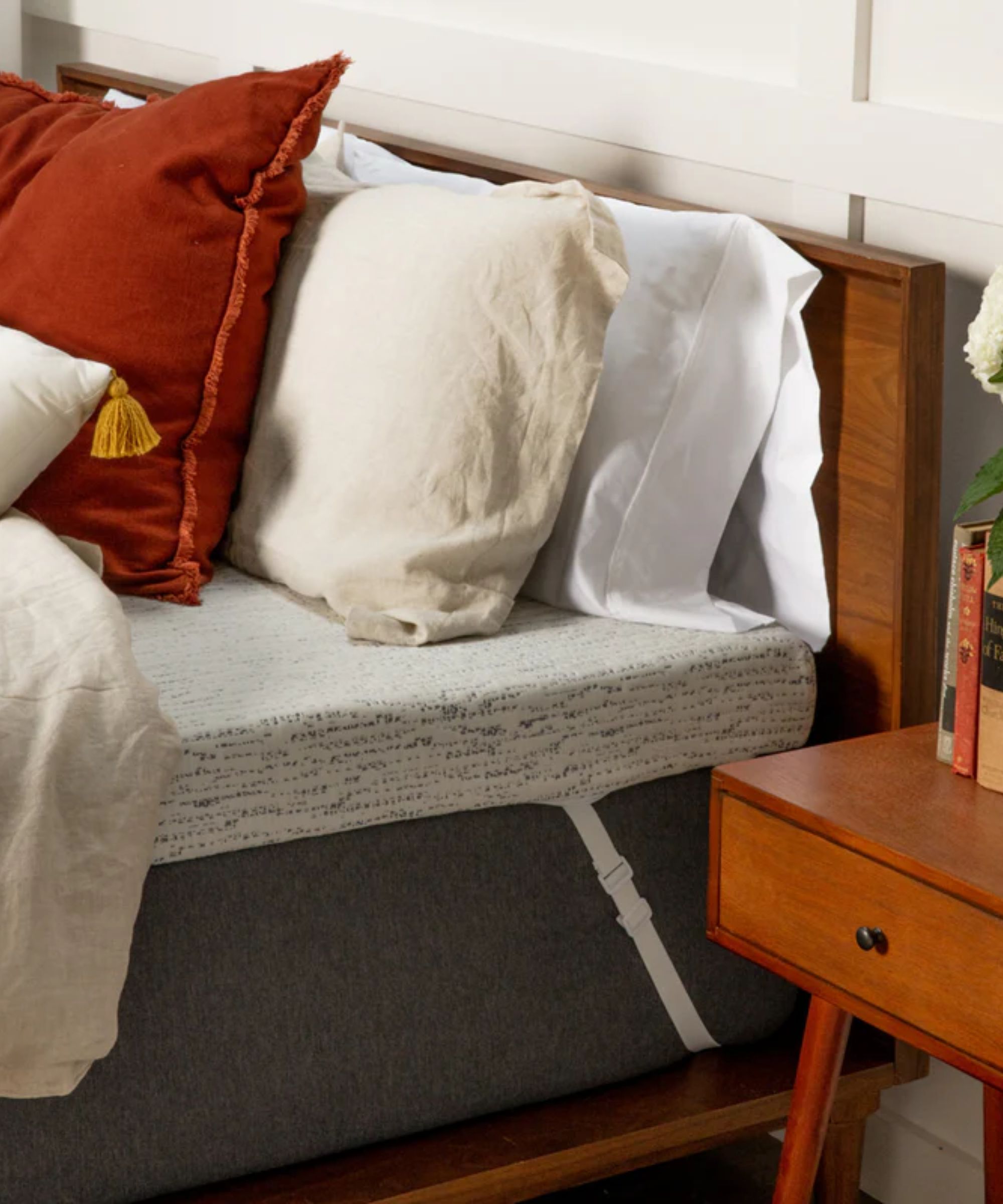 A grey Viscosoft High-Density Mattress Topper with corner straps seen on a grey mattress on a wooden bedframe. On top of the bed are an assortment of red, cream and white pillows and cushions, and behind the bed is a white panelled wall. To the right of the bed is a brown wooden bedside table with a row of books and white flowers on top.