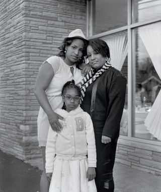 Black and white image of the Flint family, three generations of females stood together, brick wall to the left, tall glass windows with curtains to the right