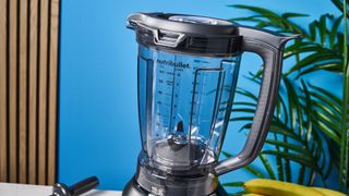 a black nutribullet with a big blending component is photographed in front of a blue backdrop, showing its plastic pitcher jug, silver blades, and tamper