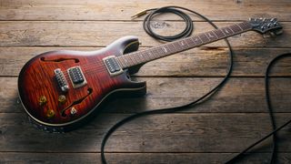 A PRS SE Ed Sheeran Hollowbody I Piezo Baritone guitar lying on a wooden floor with a guitar cable trailed nearby