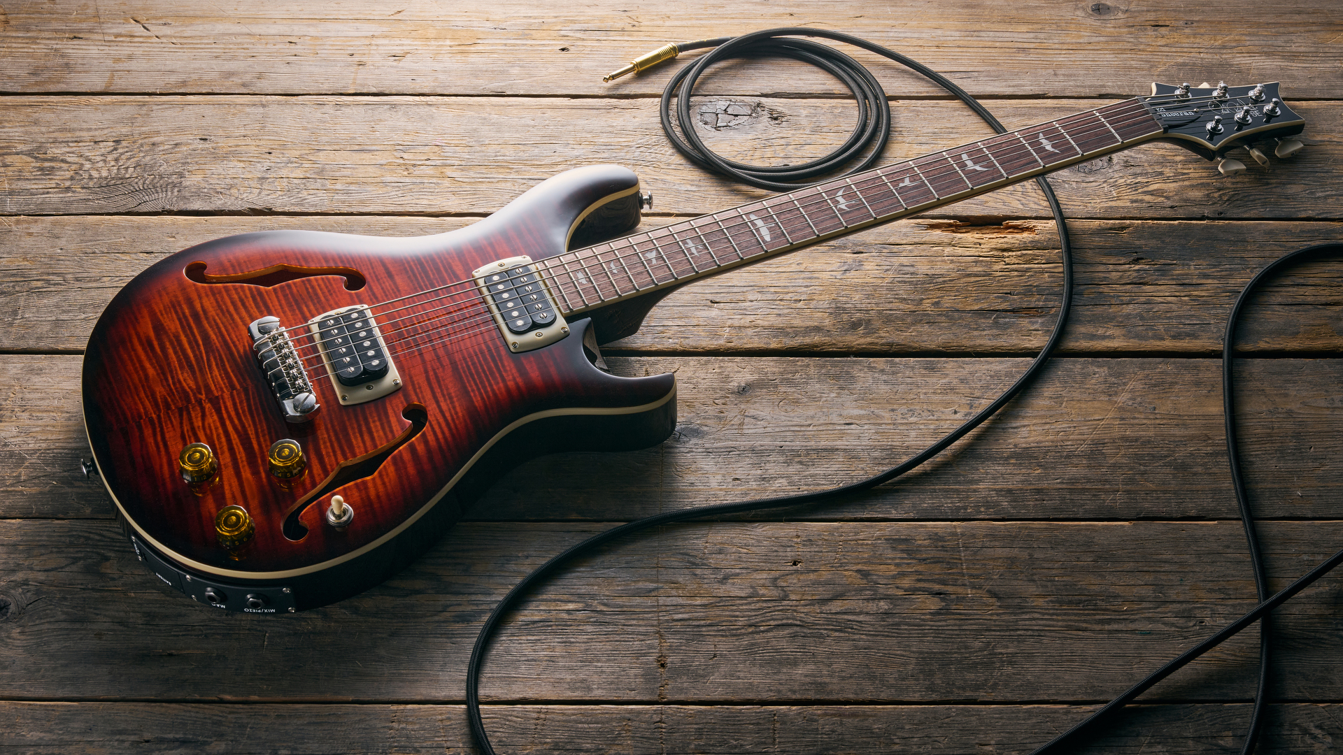 A PRS SE Ed Sheeran Hollowbody I Piezo Baritone guitar lying on a wooden floor with a guitar cable trailed nearby