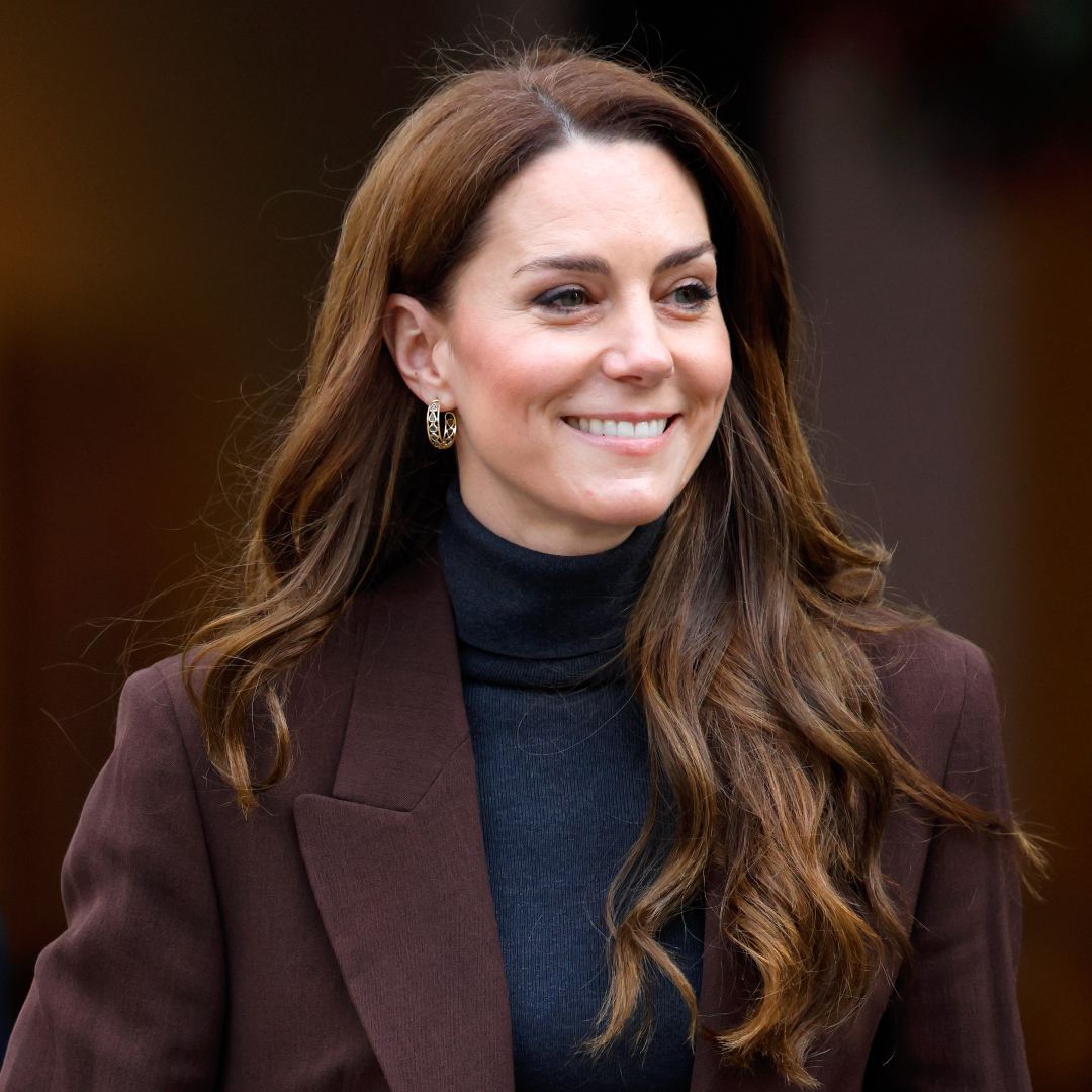 Princess Kate Middleton has long wavy brown hair and wears a black polo neck sweater with a brown blazer