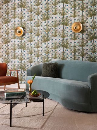 Living room with palm patterned wallpaper, brass sconces, blue velvet curved sofa and black glass coffee table