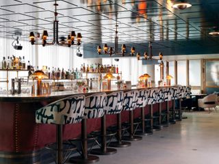 Interior view of the main bar at White City House featuring a bar with a maroon coloured front and dark brown top, multicoloured patterned bar stools, a dark coloured panelled ceiling, multi arm pendant lights, lamps and shelving with multiple drinks behind the bar