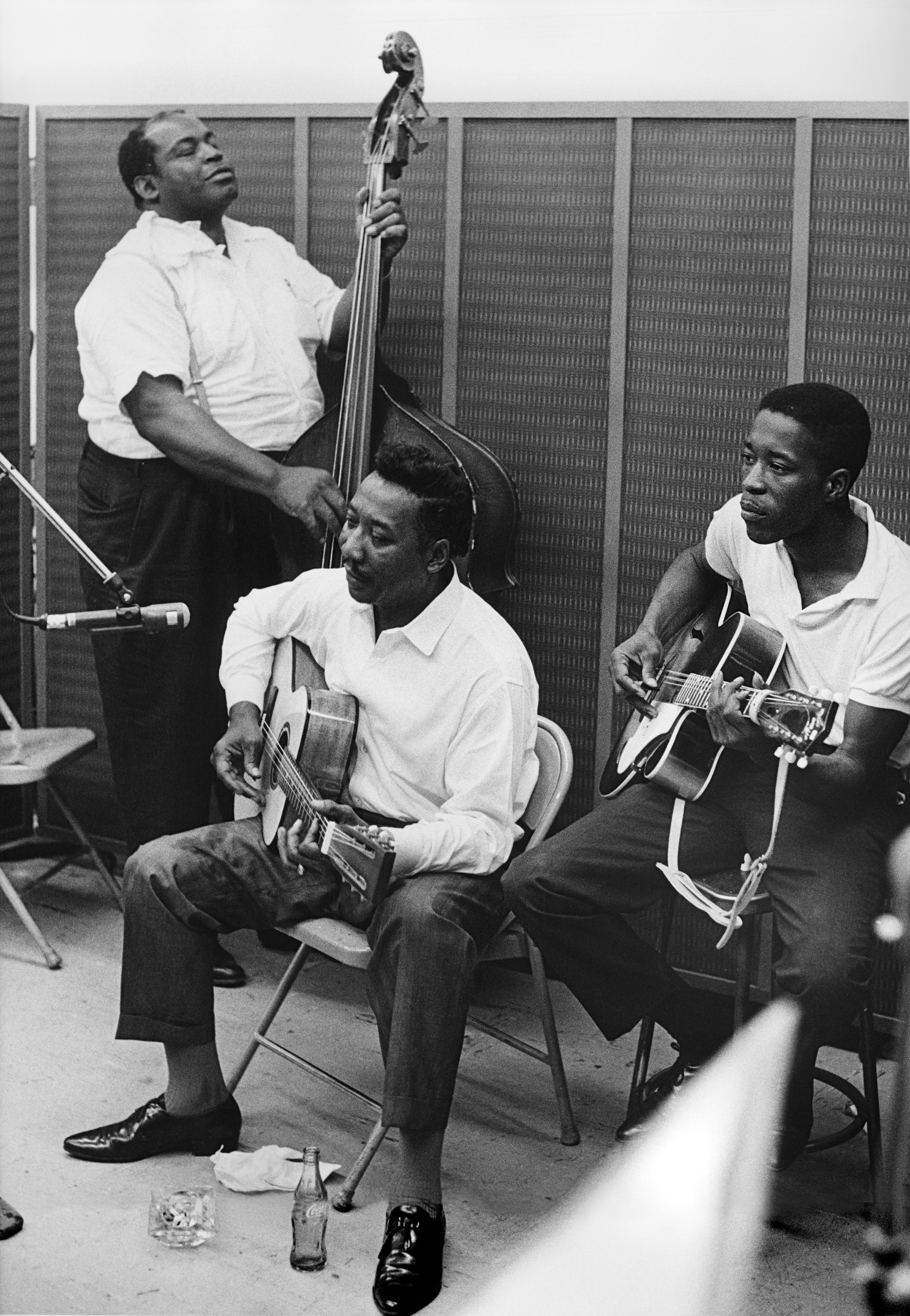A black and white candid photo of Muddy Waters and Buddy Guy playing acoustic guitars while Willie Dixon plays the upright bass behind them.