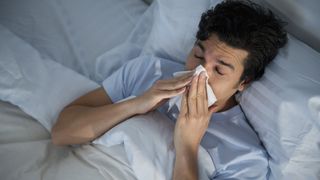 A man lies in bed blowing his nose with his head propped up on pillows to help him sleep with allergies
