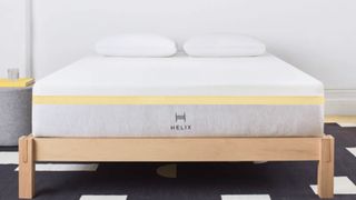 The image shows the Helix Dawn mattress on a wooden bed frame against a white wall and on a geometrical rug