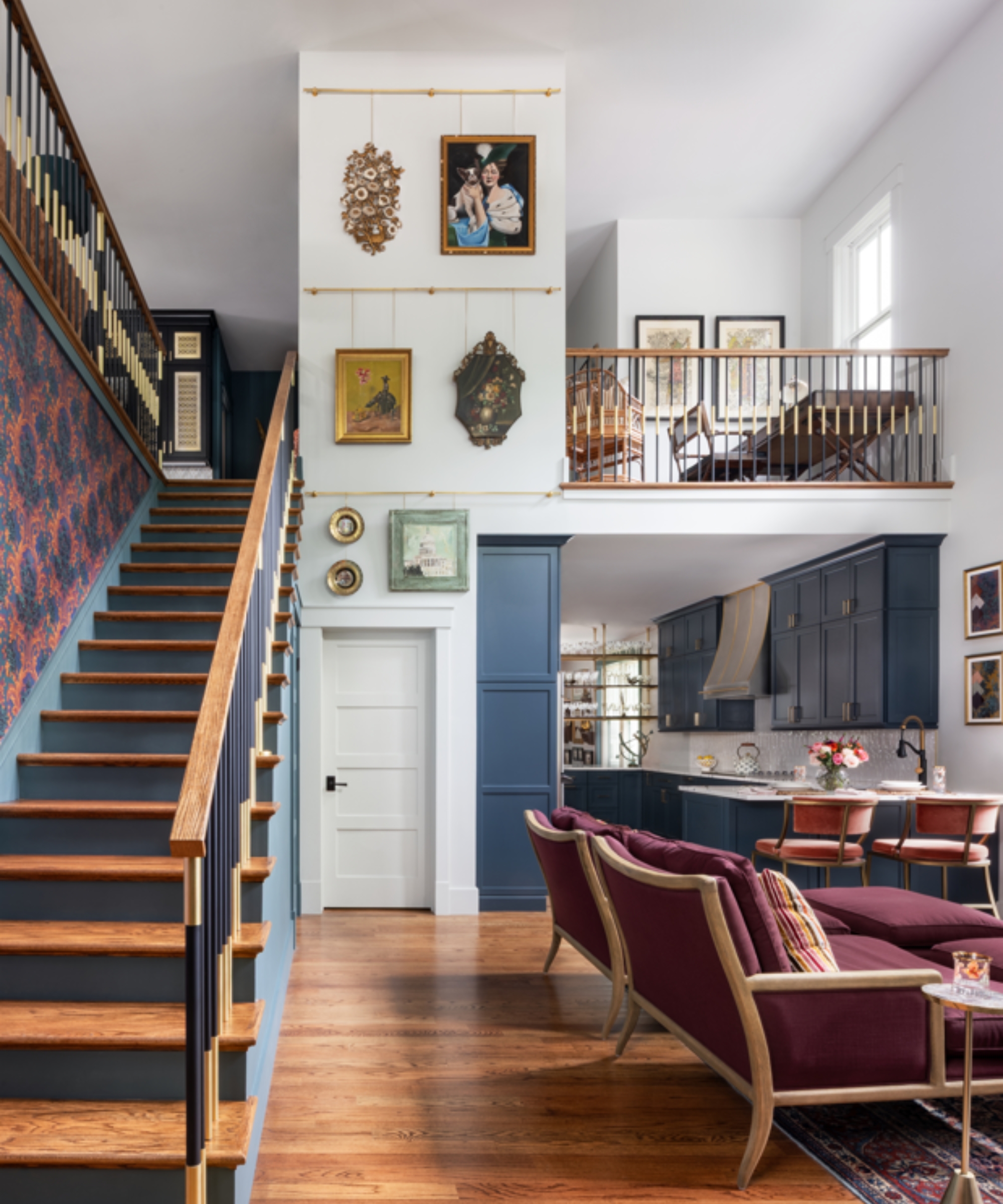 Upstairs-downstairs shot of home interior with vibrant art on white walls, wooden floors, large red sofa, and a view of an upper seating area and downstairs kitchen in background.