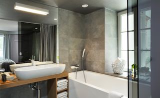 Bathroom, stone wall tiles, white bath with white shower head and silver chord, window, mesh grey divider, wooden unit with white sink on top, wall mirror behind sink, strobe light above sink, spotlight in ceiling, white towels