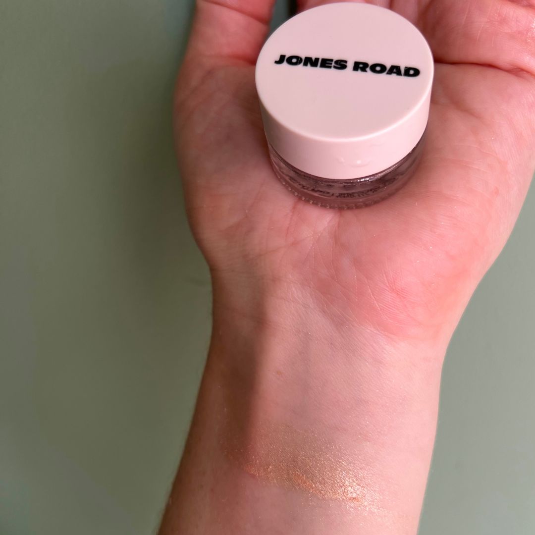 a hand on a green background holding x cream eyeshadow with a swatch of the eyeshadow on the wrist 