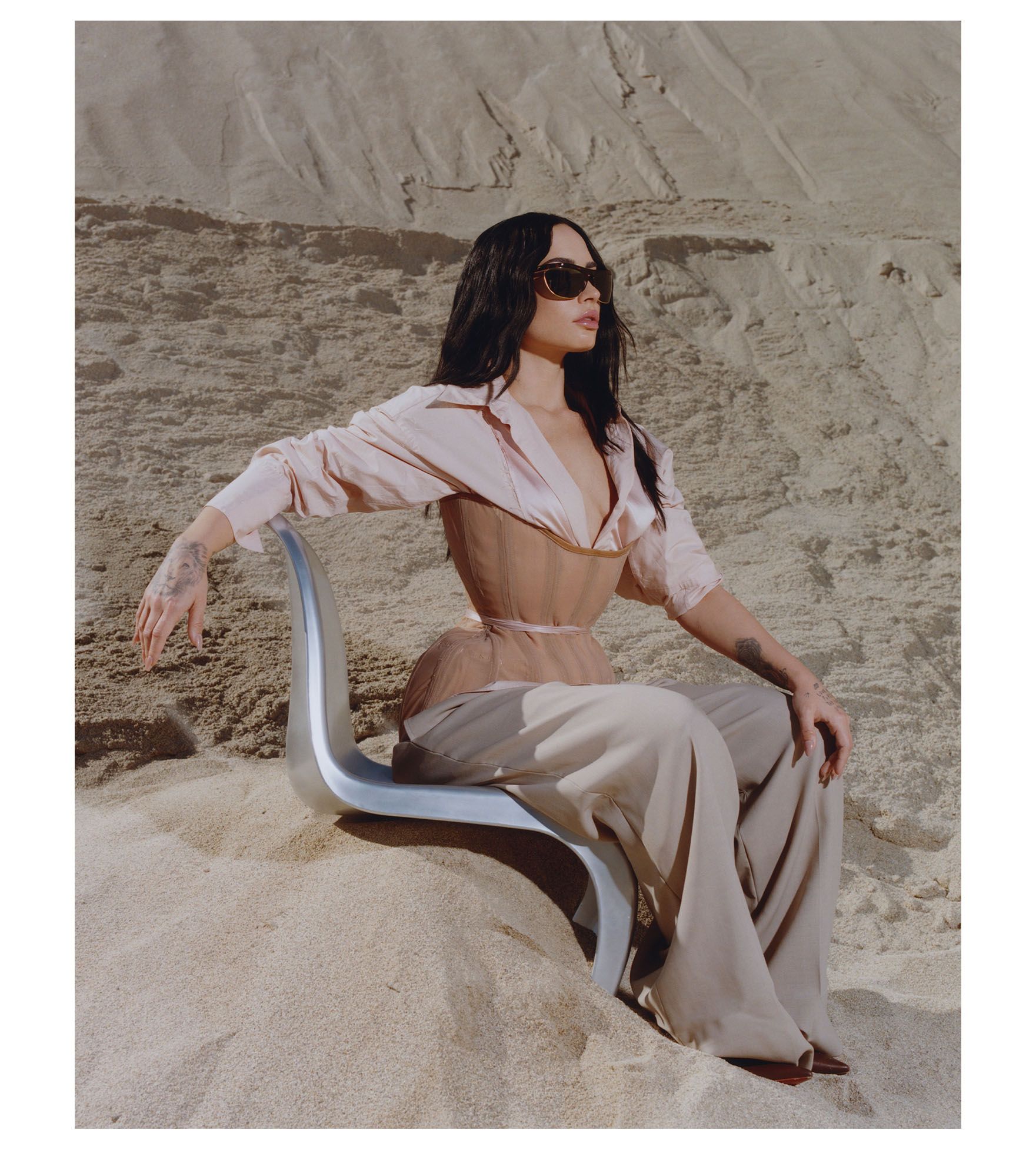 Demi Lovato for Who What Wear's November cover. She is sitting on a silver chair with her left hand on her knee on a sand dune. She is wearing a pink button-down shirt with a nude corset on top and tan trousers. She is also wearing sunglasses.