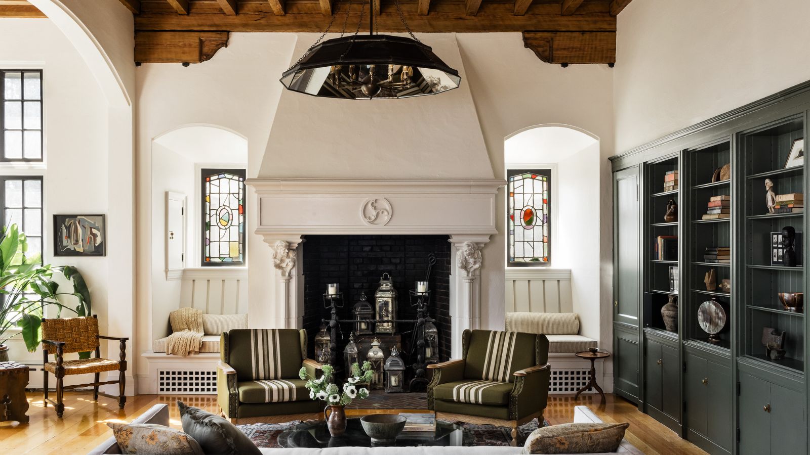 Textured living room with wooden-beamed ceiling, dark green bookshelves to the right, two striped armchairs, stained glass windows, a hanging lightshade, and wooden floors.