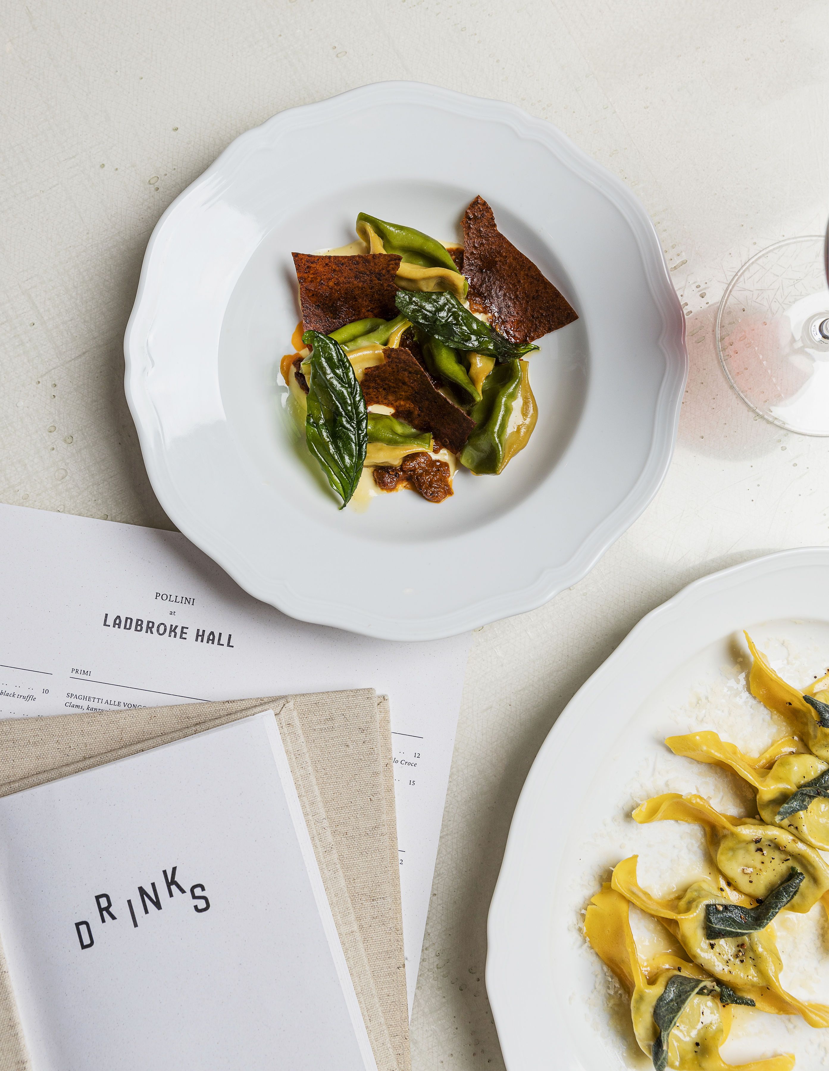 Two plated dishes of handmade pasta served in beautiful compositions on a white linen cloth next to a menu that reads "LADBROKE HALL" and "DRINK".