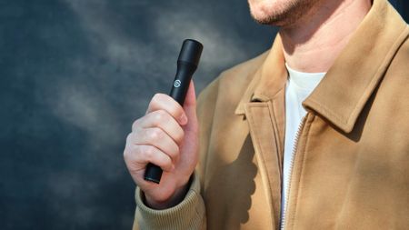 A man in a tan jacket holds a black vape pen near his chest against a dark, smoky backdrop, creating a moody, dramatic impression.