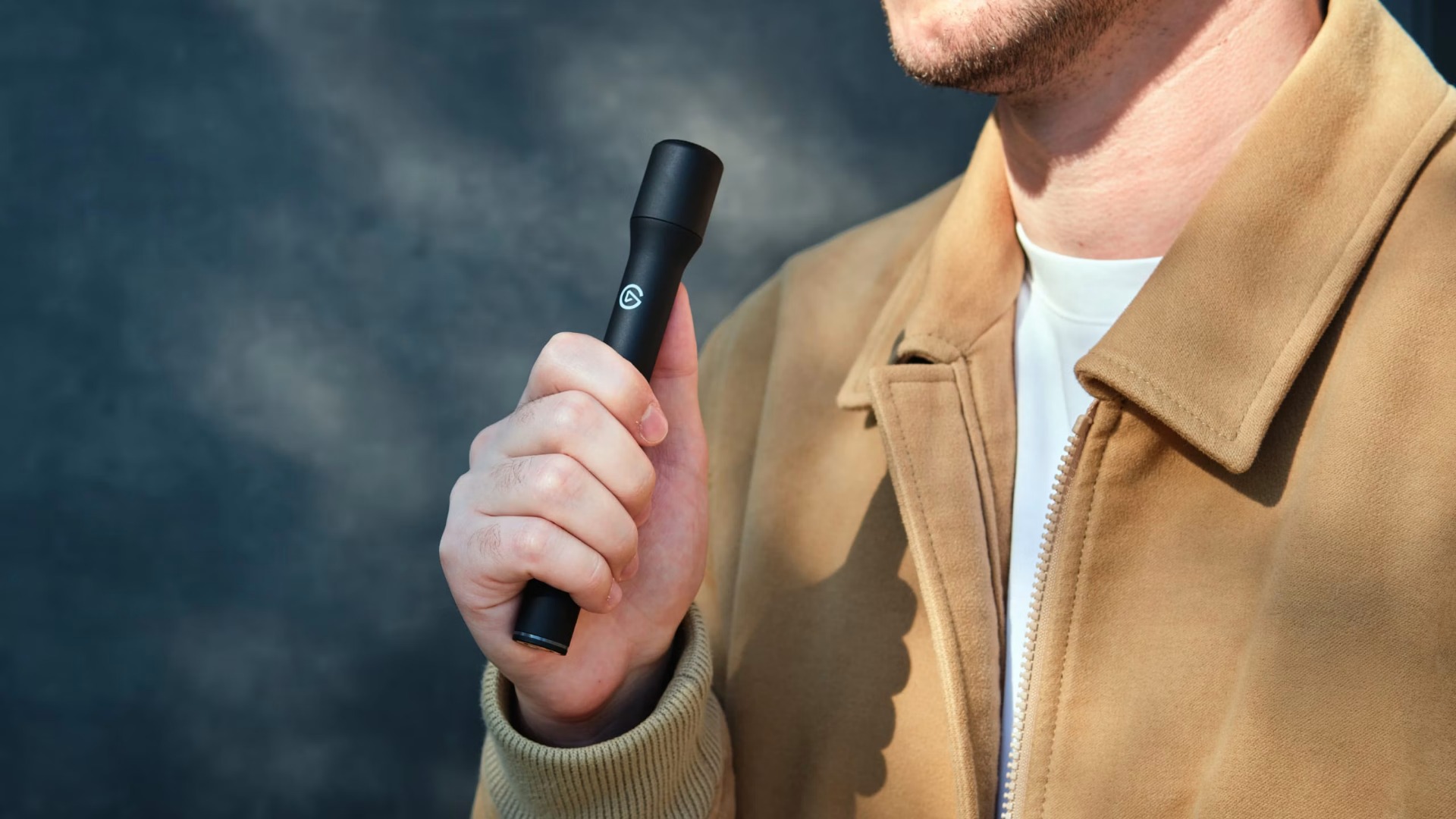 A man in a tan jacket holds a black vape pen near his chest against a dark, smoky backdrop, creating a moody, dramatic impression.